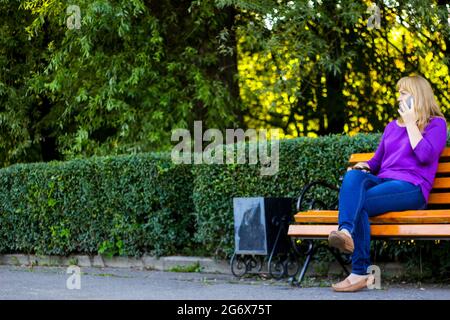 Flou artistique Femme blonde caucasienne parlant, parlant au téléphone à l'extérieur, à l'extérieur. femme de 40 ans dans un chemisier violet dans le parc. Femme adulte mobile Banque D'Images