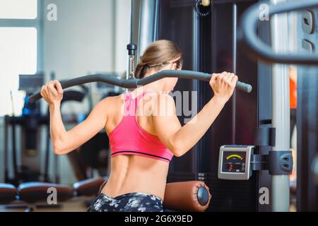 Vue arrière d'une femme en forme de forte exerçant un mouvement de pression latéral derrière le cou au niveau de la machine à câble pour les muscles du dos dans un club de fitness moderne Banque D'Images