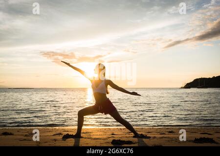 Vue latérale sur toute la longueur de la silhouette d'une femme fit pratiquer le yoga guerrier contre le ciel au coucher du soleil pendant les vacances d'été dans l'île de Flores, Banque D'Images