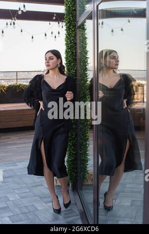 pleine longueur de jeune femme en robe noire et veste en fausse fourrure debout avec la main sur la hanche sur la terrasse Banque D'Images