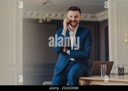 Photo de beau barbu homme a la conversation téléphonique rend l'appel cellulaire satisfait avec de bonnes poses de nouvelles au bureau avec ordinateur portable moderne Banque D'Images