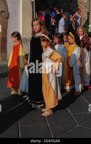 Des jeunes filles et un garçon tout-petit, tous en costume médiéval coloré, tiennent les mains et bavardez pendant qu'ils attendent à Piazza Guglielmo Marconi à Anagni, Lazio, Italie, pour un signal de marcher dans un pageant historique d'août en l'honneur de Saint Magnus d'Anagni, leur Saint patron. Le spectacle rappelle également le passé d'Anagni en tant que ville des Papes, la résidence d'été du pontife à la recherche d'un refuge plus frais face à la chaleur estivale de Rome. Les filles portent de longs pardessus ou pardessus tandis que le petit garçon est vêtu d'une blouse blanche pour bébé. Banque D'Images