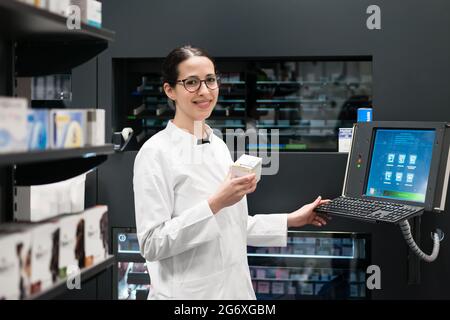 Low-angle rear view of an experienced female pharmacist using a computer while managing the drug stock in a contemporary pharmacy with modern technolo Stock Photo