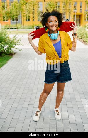 Femme afro-américaine avec casque bleu tient le skateboard entre les mains lors de la marche ville moderne profitant d'activités de loisirs d'été Banque D'Images