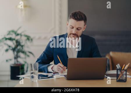 Sérieux concentré de l'entrepreneur de sexe masculin vérifie rapport financier de comptabilité écrit quelques notes dans le bloc-notes se trouve dans l'espace de collègue utilise ordinateur portable Banque D'Images