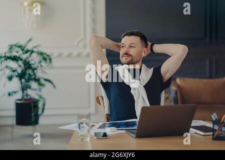 Un homme européen expérimenté repose sur une chaise et regarde loin avec des poses d'expression réfléchis sur des poses de bureau au bureau à domicile écoute les informations via des écouteurs Banque D'Images