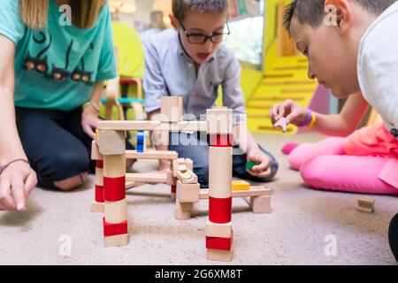 Vue à angle bas d'un enseignant de jardin d'enfants dédié aidant les enfants avec la construction d'un circuit de train en bois pendant les temps de jeu libre supervisé Banque D'Images