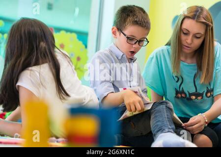 L'école pré-cute boy wearing eyeglasses tandis que l'apprentissage de la lecture d'un livre pour enfants aidés par un assistant enseignant de maternelle Banque D'Images