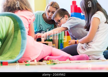 Cute pre-school boy cooperating with his colleagues at the construction of a structure, made of wooden toy blocks under the guidance of a young kinder Stock Photo