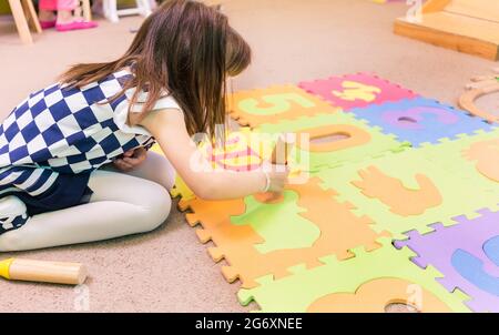 Vue latérale d'une adorable fille d'avant-école tenant des blocs de jouets en bois, tout en jouant avec un tapis de jeu de puzzle coloré avec des nombres et des signes dans la salle de classe o Banque D'Images