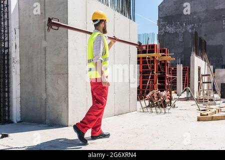 Longueur totale d'un ouvrier de col bleu portant de l'équipement de sécurité, tout en portant une lourde barre métallique pendant les travaux sur le site de construction d'une résidence Banque D'Images