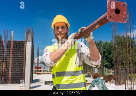 Longueur totale d'un ouvrier de col bleu, portant de l'équipement de sécurité tout en portant une lourde barre métallique pendant les travaux sur le site de construction d'une résidence Banque D'Images