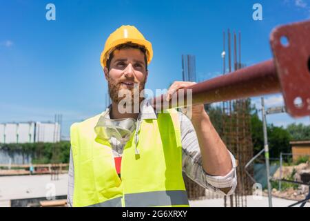 Longueur totale d'un ouvrier de col bleu, portant de l'équipement de sécurité tout en portant une lourde barre métallique pendant les travaux sur le site de construction d'une résidence Banque D'Images