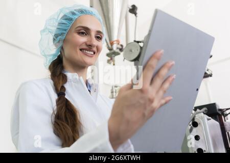 Expert femelle portant sarrau blanc et casque de protection lors de l'analyse de l'information sur un tablet PC pendant le travail dans une usine contemporaine Banque D'Images