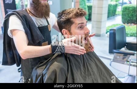 Vue latérale d'un jeune homme barbu à tête rouge faisant un visage amusant, tout en étant assis sur la chaise coiffante avant un changement de look au salon de coiffure Banque D'Images