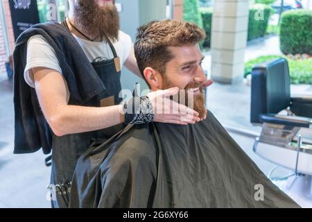 Beau jeune homme assis sur une chaise coiffante, tout en souriant et pensant à une forme différente pour sa barbe sur les conseils de son coiffeur dans le bea Banque D'Images