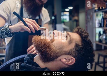 Vue latérale : tête d'un jeune homme barbu à tête rouge souriant prêt à se raser dans le salon de coiffure, d'un coiffeur qualifié avec un rasoir droit classique en haut Banque D'Images