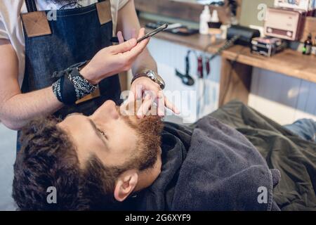 Vue latérale d'un tir à la tête rousse jeune homme barbu souriant, prêt pour le rasage dans le salon de coiffure d'une main-d'coiffure avec un rasoir droit classique en hi Banque D'Images