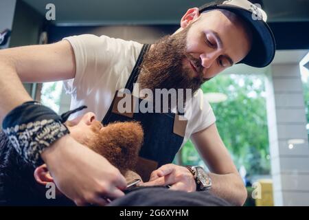 Vue latérale d'un tir à la tête rousse jeune homme barbu souriant, prêt pour le rasage dans le salon de coiffure d'une main-d'coiffure avec un rasoir droit classique en hi Banque D'Images