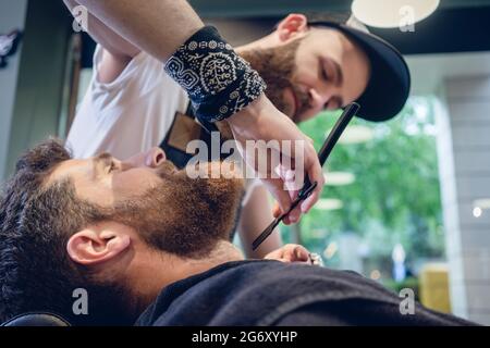 Vue latérale d'un tir à la tête rousse jeune homme barbu souriant, prêt pour le rasage dans le salon de coiffure d'une main-d'coiffure avec un rasoir droit classique en hi Banque D'Images