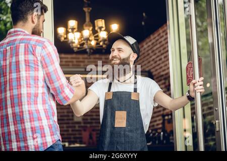 Vue en contre-plongée d'un coiffeur gai avec une barbe hipster, portant un tablier tout en saluant son client mâle à l'entrée d'un salon de coiffure cool pour hommes Banque D'Images