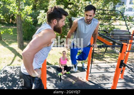 Deux jeunes hommes musclés faisant des exercices de poids corporel pour la partie supérieure du corps-motivés par leur amie dans un parc de remise en forme moderne en été Banque D'Images