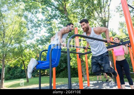 Deux jeunes hommes forts et motivés faisant des exercices de creux pour le haut du corps, pendant l'entraînement stimulant avec leur ami dans un parc de fitness extérieur avec esprit Banque D'Images