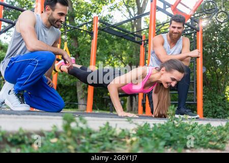 Vue en contre-plongée d'une jeune femme en forme faisant un exercice d'extension de jambe avec une suspension d'entraînement, motivée par ses amis sportifs joyeux dans un calis moderne Banque D'Images