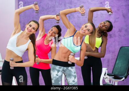 Quatre femmes gaies tenant des haltères tout en faisant des exercices pour les bras et les muscles abdominaux latéraux, pendant le cours de fitness de groupe dans un club de santé moderne Banque D'Images