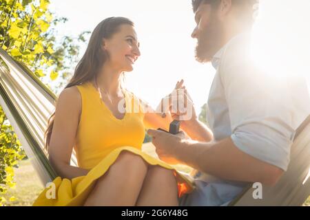 Vue en contre-plongée d'un jeune homme heureux mettant un anneau d'engagement sur le doigt de sa petite amie, après avoir accepté sa proposition tout en étant assis ensemble Banque D'Images