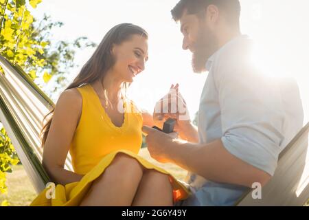 Vue en contre-plongée d'un jeune homme heureux mettant un anneau d'engagement sur le doigt de sa petite amie, après avoir accepté sa proposition tout en étant assis ensemble Banque D'Images