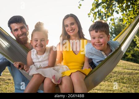 Mignon enfant faisant un visage drôle, tout en regardant l'appareil photo pour un portrait de famille avec sa sœur et ses parents sur un hamac à l'extérieur dans une journée ensoleillée de summ Banque D'Images
