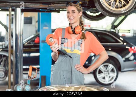 Portrait d'une femme gaie mécanicien automobile portant des équipements de sécurité tout en tenant une machine à polir de voiture dans un atelier de réparation automobile moderne Banque D'Images