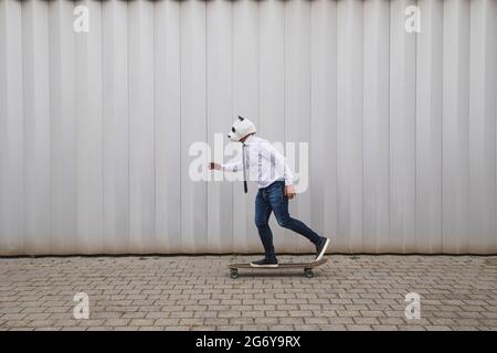 Un homme d'affaires fait du skateboard sur une longue planche avec un masque d'ours en panda contre un mur gris Banque D'Images