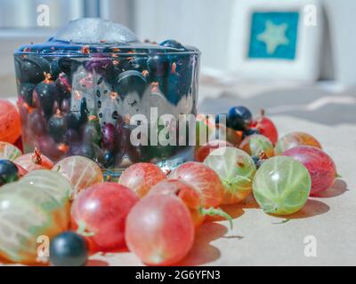 Baies de cassis dans un verre d'eau avec le cube de glace. Les groseilles à maquereau colorées sont dispersées autour de la tasse. Fenêtre et coque en arrière-plan. Banque D'Images