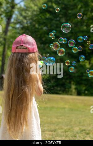 Petite fille avec un chapeau rose et de longs cheveux soufflant des bulles de savon dans l'air devant le fond de la forêt. Fille debout à l'appareil photo Banque D'Images