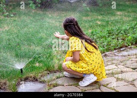 vue arrière d'une fille qui joue avec des gouttes d'eau d'un arroseur de pelouse, une fille aide ses parents à arroser l'herbe sur la pelouse Banque D'Images