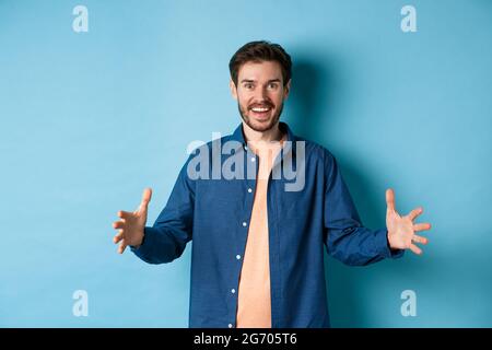 Joyeux homme souriant montrant un objet de grande taille, façonnant de grandes choses et regardant stupéfait, debout sur fond bleu Banque D'Images