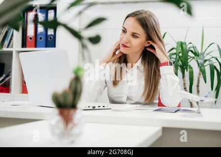 Femme d'affaires détendue utilisant un ordinateur portable et un smartphone pour communiquer et travailler en ligne dans un environnement moderne avec des plantes. L'agence de voyage a du mal à faire Banque D'Images