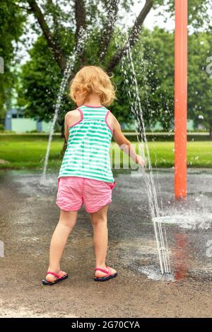 Un petit enfant joue avec de l'eau dans le parc public local le jour d'été chaud. Petite belle fille s'amuser à la fontaine de jeux. Banque D'Images