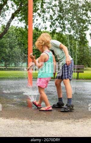 Des enfants heureux jouent avec de l'eau dans le parc public local le jour d'été chaud et ensoleillé. Les petits enfants s'amusent à la fontaine Banque D'Images