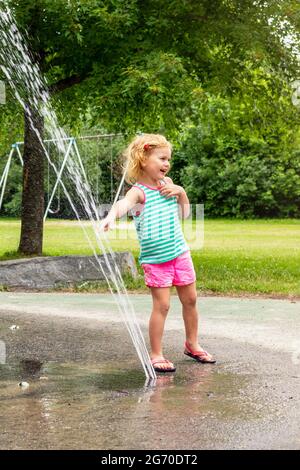 Un petit enfant souriant joue avec de l'eau dans le parc public local le jour d'été chaud. Petite belle fille s'amuser à la fontaine Playgrou Banque D'Images