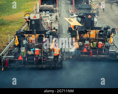 Plusieurs ouvriers avec des casques de sécurité et des gilets orange réfléchissants faisant fonctionner des machines de pavage d'asphalte CAT et des camions à benne basculante sur l'autoroute 25 au Québec. Banque D'Images