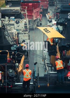 Plusieurs ouvriers avec des casques de sécurité et des gilets orange réfléchissants faisant fonctionner des machines de pavage d'asphalte CAT et des camions à benne basculante sur l'autoroute 25 au Québec. Banque D'Images