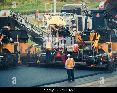 Plusieurs ouvriers avec des casques de sécurité et des gilets orange réfléchissants faisant fonctionner des machines de pavage d'asphalte CAT et des camions à benne basculante sur l'autoroute 25 au Québec. Banque D'Images