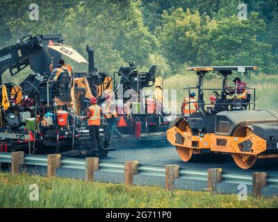 Plusieurs ouvriers avec des casques de sécurité et des gilets orange réfléchissants faisant fonctionner des machines de pavage d'asphalte CAT et des camions à benne basculante sur l'autoroute 25 au Québec. Banque D'Images