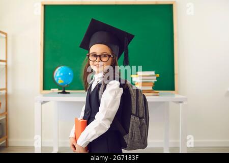 Petite fille intelligente dans le chapeau de mortier de graduation posant à la salle de classe. Enfant intelligent avec sac à dos le premier jour de l'école Banque D'Images