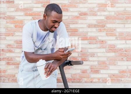 afro-américain jeune homme reposant le bras sur son scooter et utilisant son téléphone. Il est les rues près d'un mur de briques. Banque D'Images