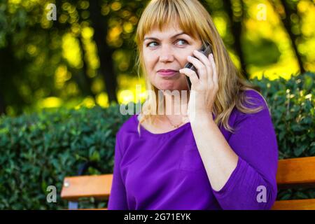 Flou artistique Femme blonde caucasienne parlant au téléphone à l'extérieur, à l'extérieur. femme de 40 ans en chemisier violet dans le parc sur banc. Une personne adorable utilisant un téléphone mobile, Banque D'Images