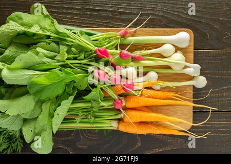 Jeunes radis et carottes fraîchement cueillis avec des sommets verts et des oignons verts sur une planche sur fond de bois blanc Banque D'Images